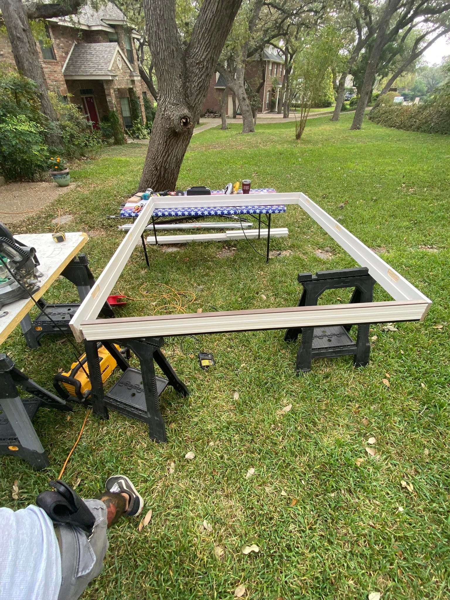 A wood frame on sawhorses in a grassy yard, near a table and saw. A person is nearby.