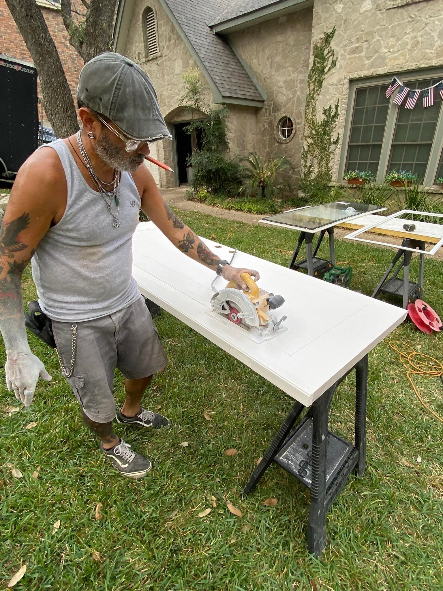 Man cuts a white board with a circular saw in a yard, near a stone house.