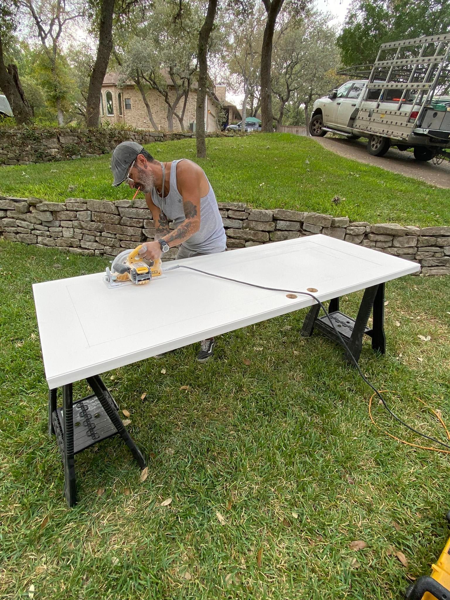 Man using a saw to cut a white board on sawhorses outdoors, near a vehicle and stone wall.
