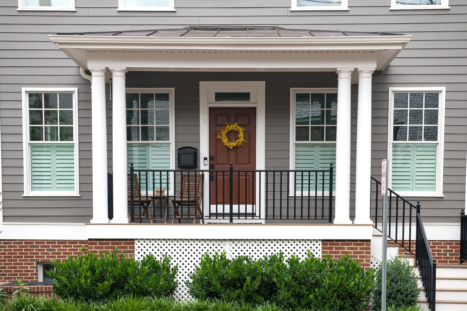 Gray house with covered porch, white columns, brown door with wreath, windows with shutters, and black railing.