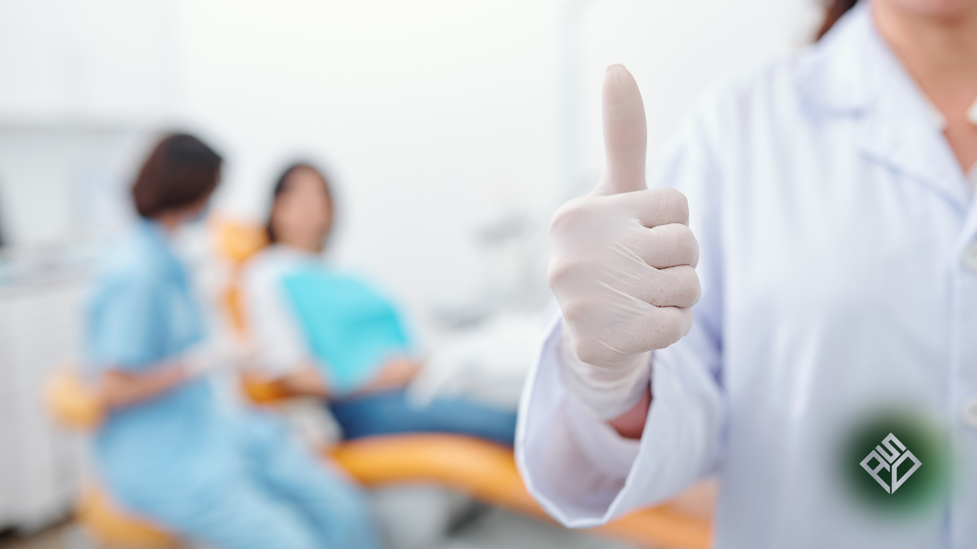 Dentist giving thumbs up in dental office. Patient and assistant in background.