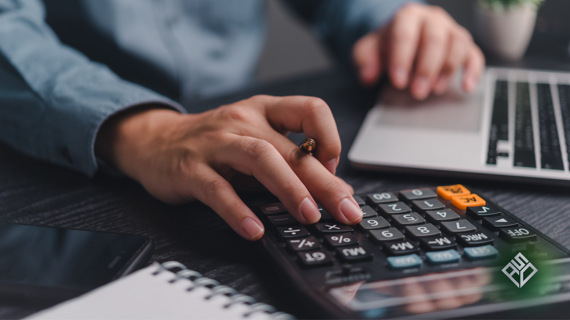Person's hands using a calculator next to a laptop and notepad, working on finances.