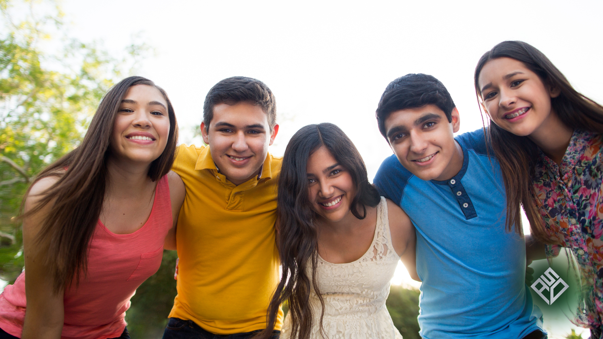 Four smiling people huddle together under a blue sky, arms around each other, looking down.