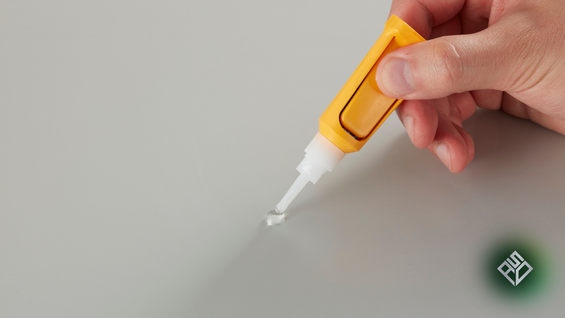 Hand applying glue from an orange tube to a gray surface.