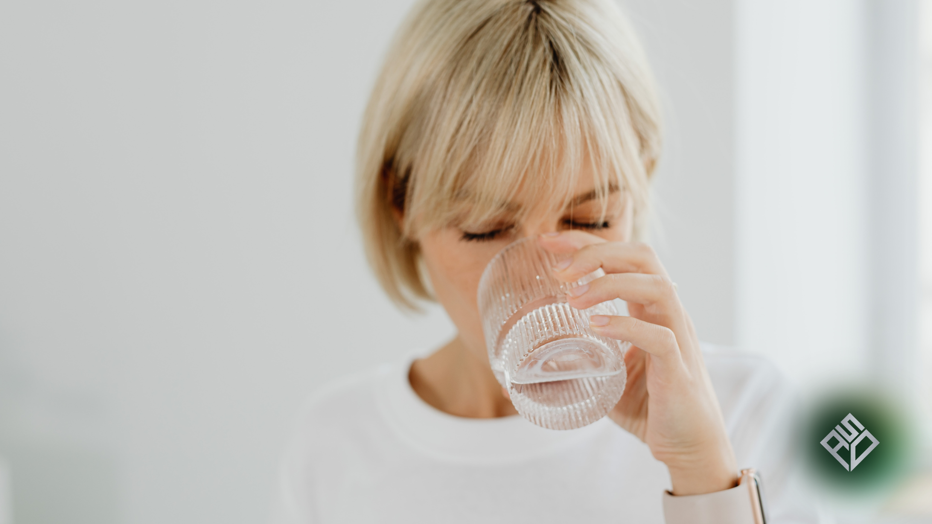 Woman drinking water from a clear glass, eyes closed, blonde hair, white top, bright setting.