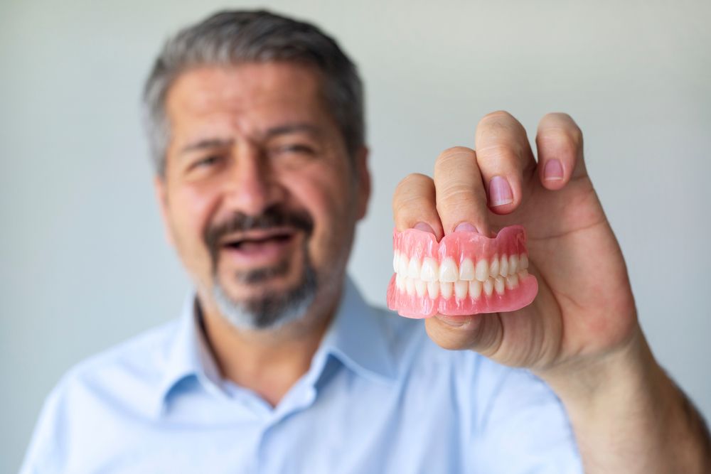 Man holds dentures, looking distressed. Gray hair, blue shirt.