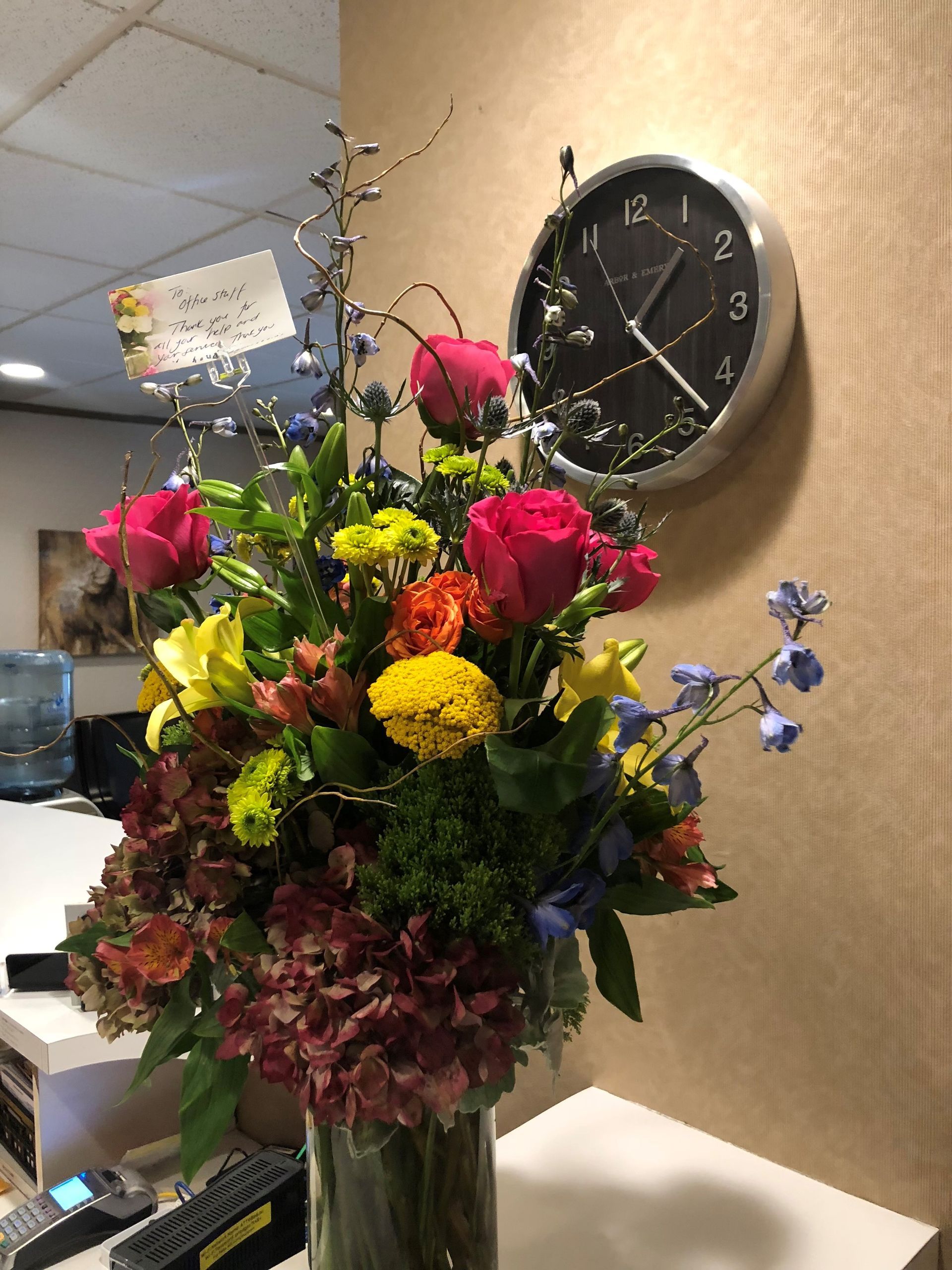 Colorful floral arrangement in a glass vase on a white surface next to a clock.