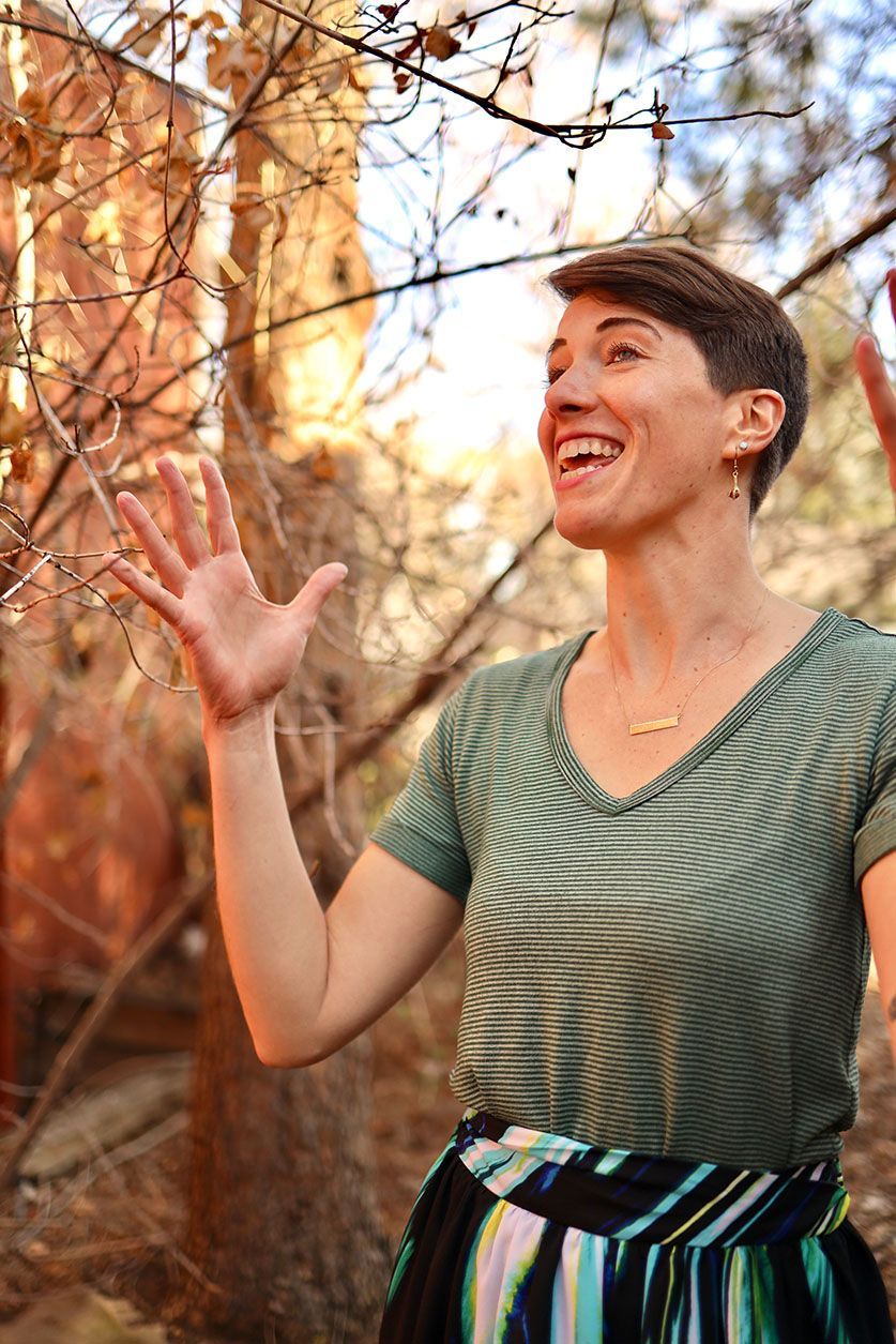 A woman is standing in the woods looking up at a tree.