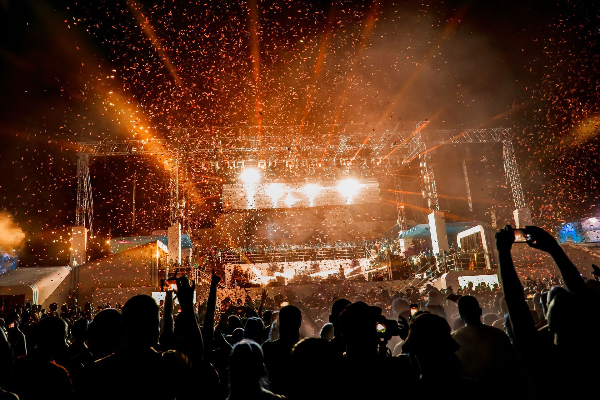 Crowd at a concert, silhouetted against a stage with bright lights, pyrotechnics, and confetti.