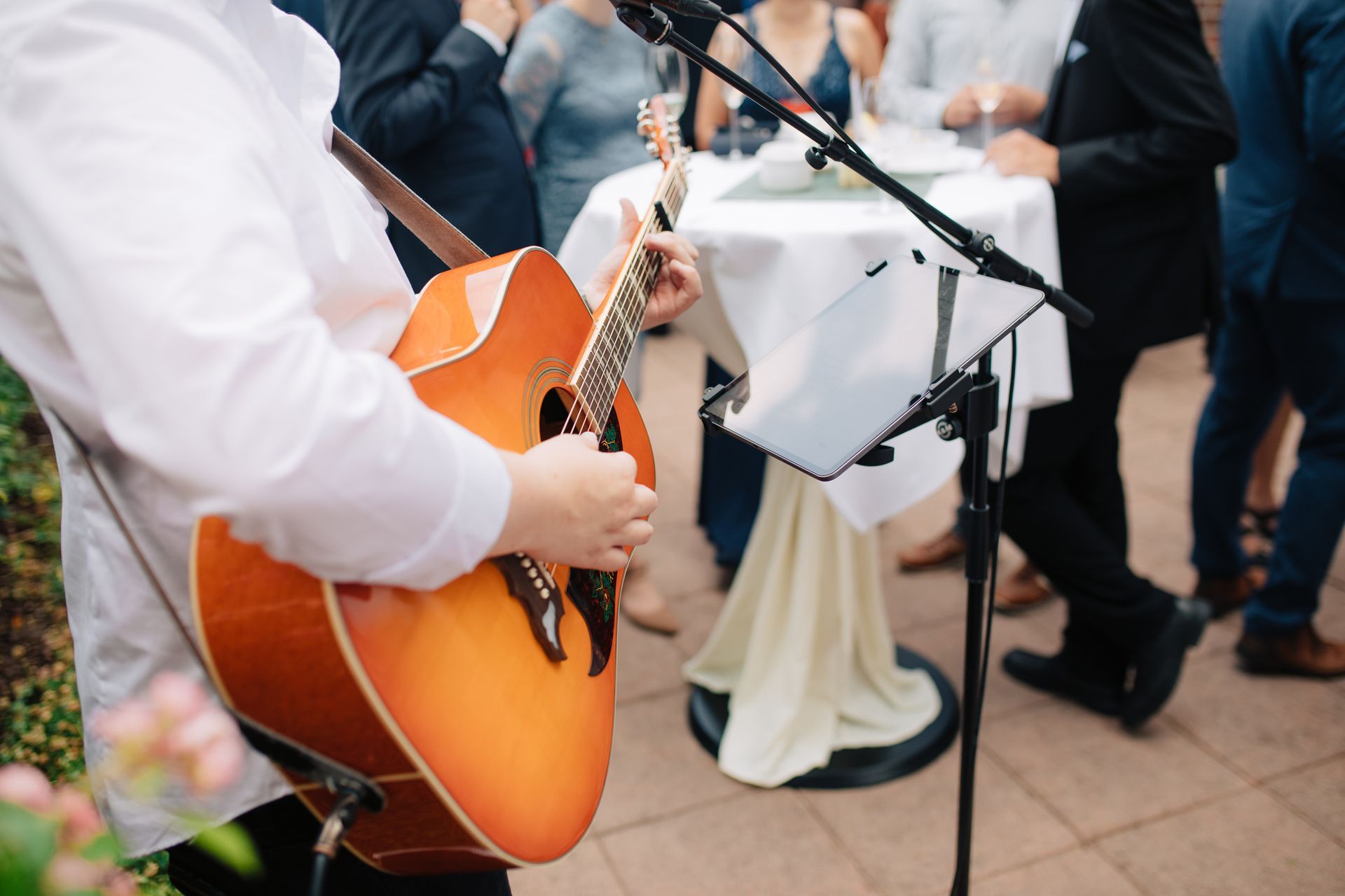 Musician playing acoustic guitar at an event. People standing nearby, white table in the background.