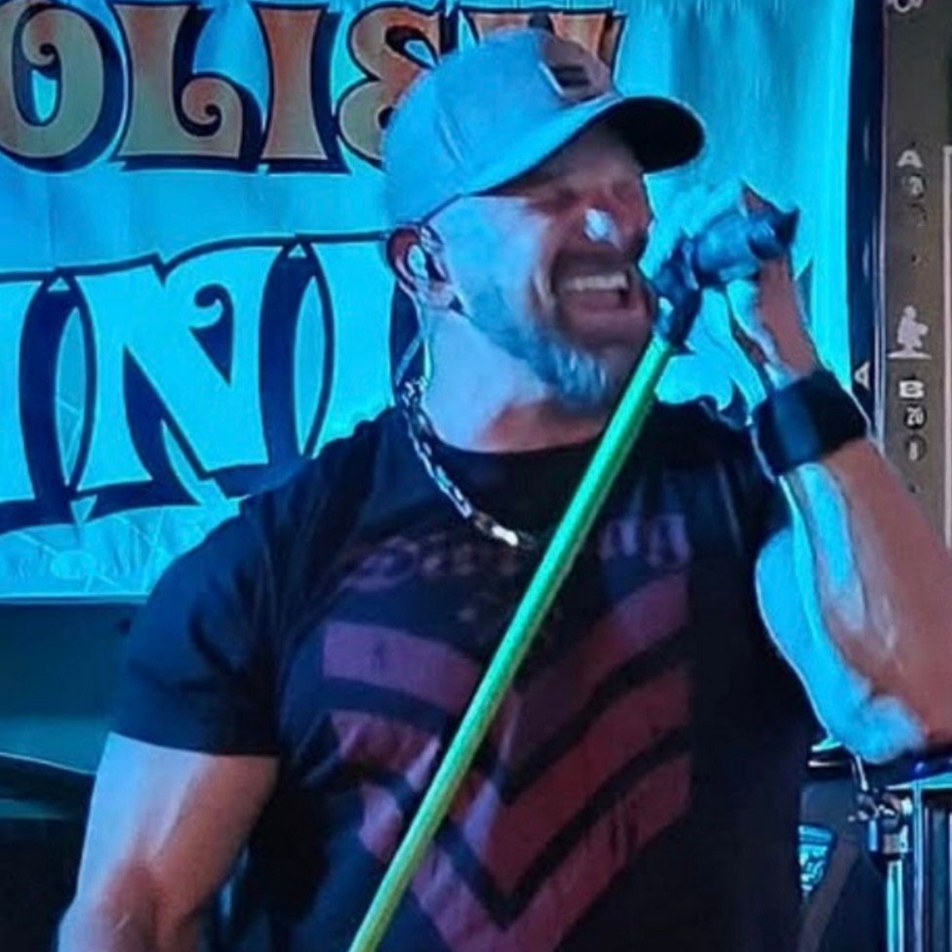 Man singing into a microphone, wearing a hat, black shirt with red stripes, and silver chain, on stage.