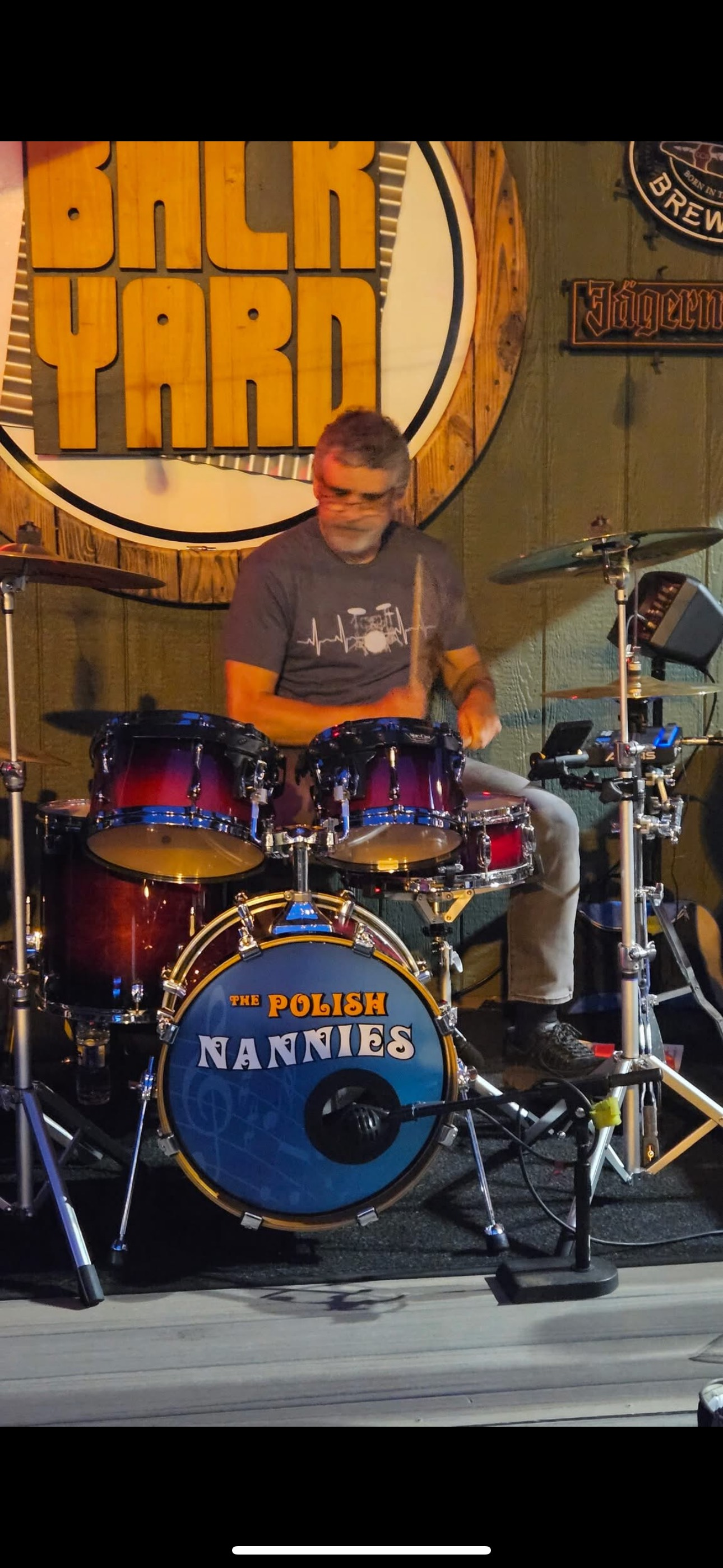 Drummer playing a drum set on stage. The drum set has a blue and red finish. A Backyard sign is in the background.