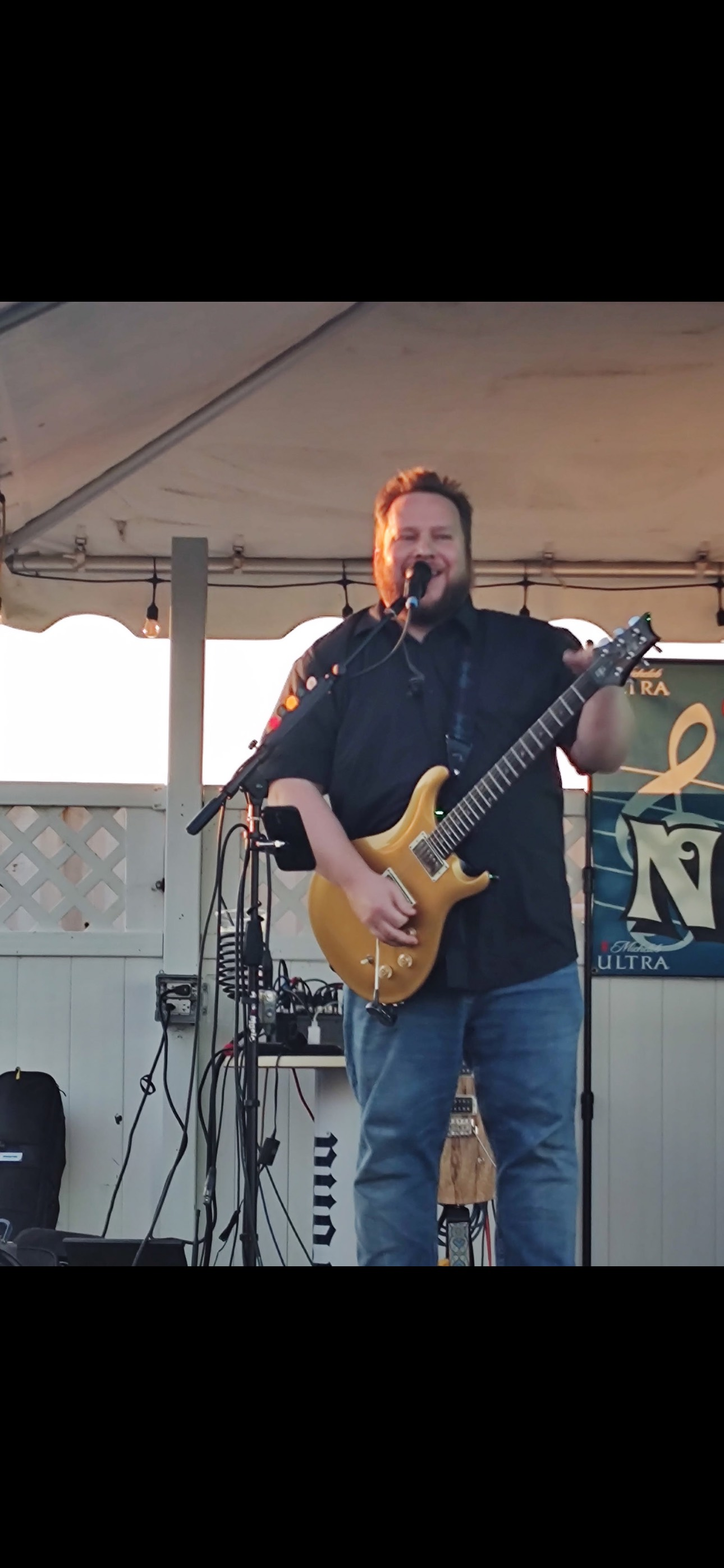 A musician plays an electric guitar on a stage under a white canopy. He's wearing blue jeans and a black shirt.