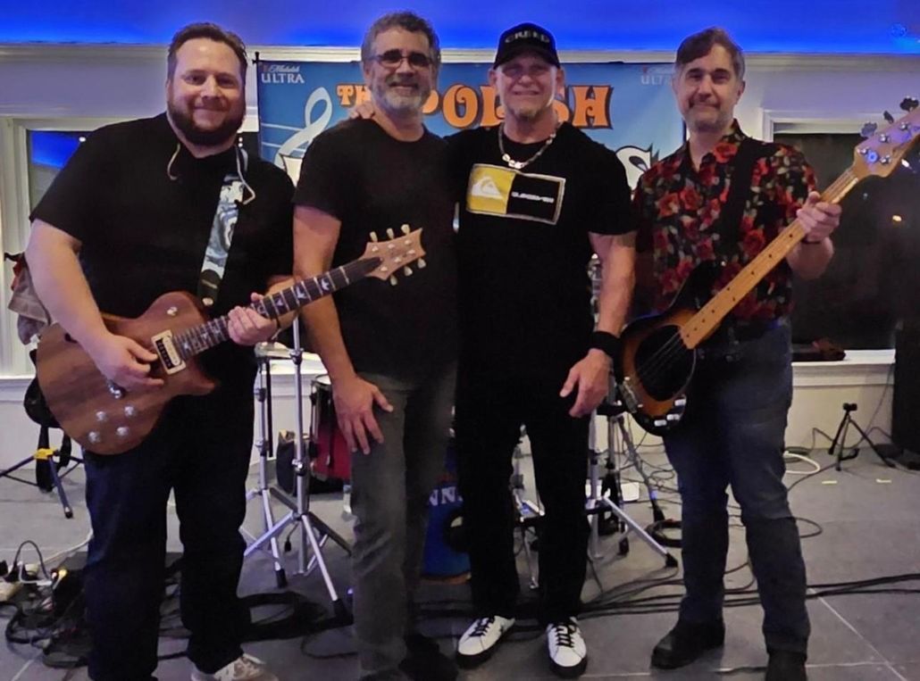 Four band members pose together: Guitar, Bass, and two men standing in between them. Band backdrop in background.