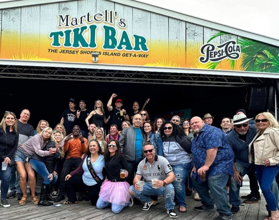 Group of people posing in front of a Tiki Bar, under a sign with a Pepsi logo.