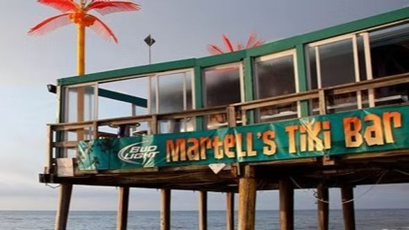 Martell's Tiki Bar on a pier with a sign, palm tree decorations, and an ocean view.