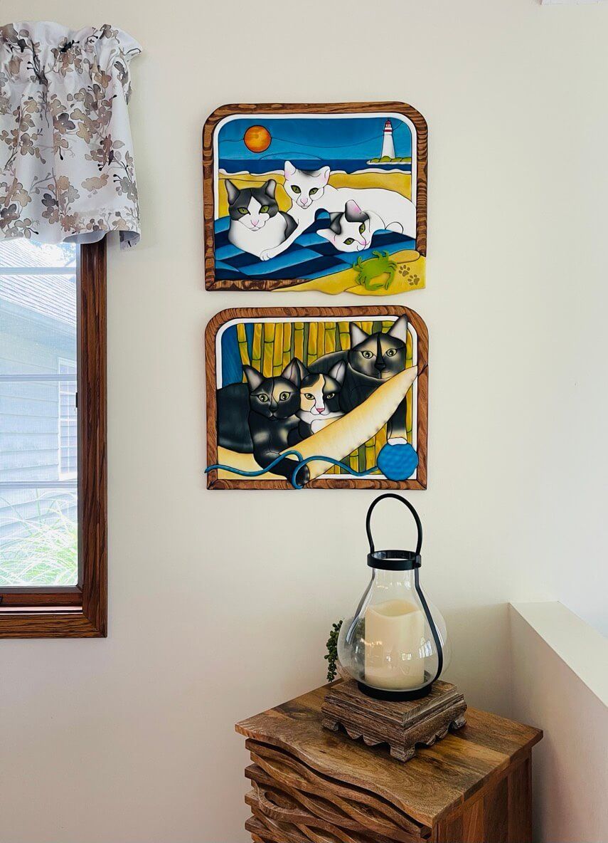 Intarsia art by Nick Vidakovic featuring two pieces with a trio of cats on a beach with lighthouse and sunset in background on beach towel with approaching crab in one scene and lying in a hammock playing with a ball of blue string in front of river reeds in the other.