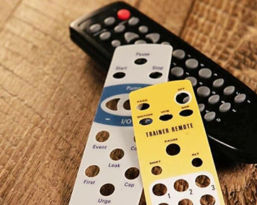 Two remote control overlays with a black remote on a wooden surface.