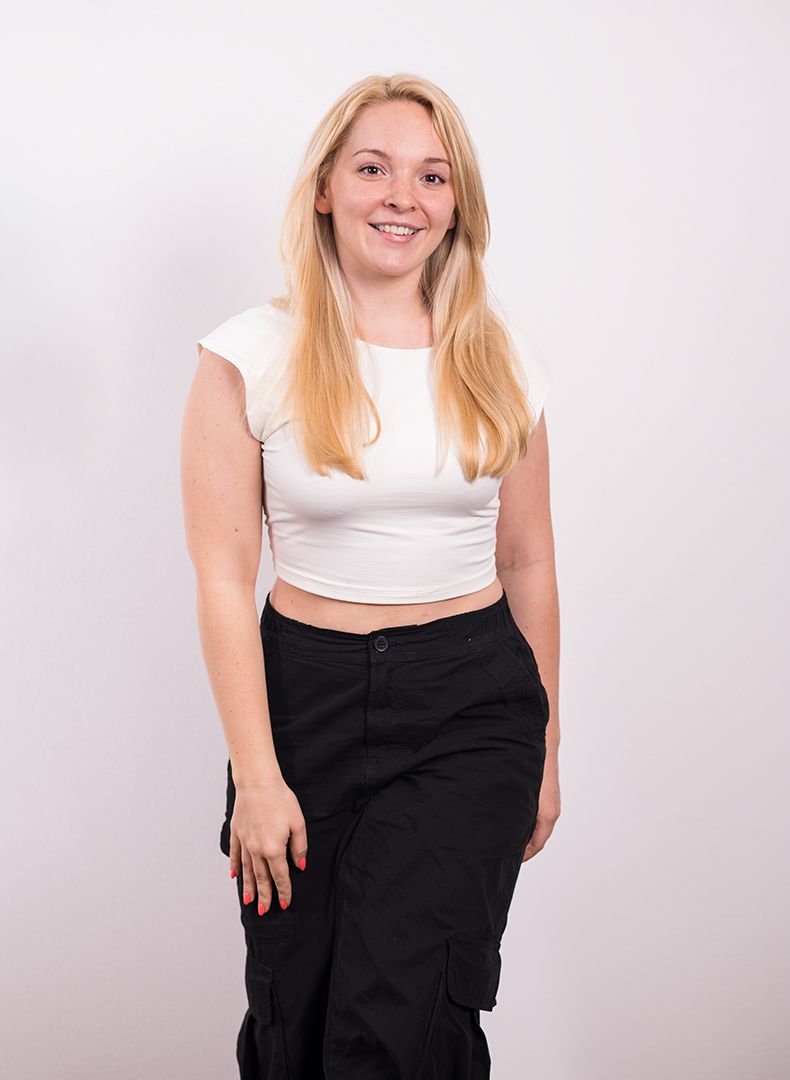 A woman in a white crop top and black pants is standing in front of a white wall.