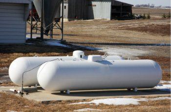 Two white propane tanks on a concrete pad, outdoors, with farm buildings in the background.