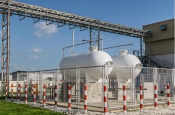 Two white cylindrical tanks inside a fenced enclosure; pipes and industrial building in the background.