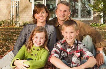 Family of four smiling, sitting outdoors in front of a house.