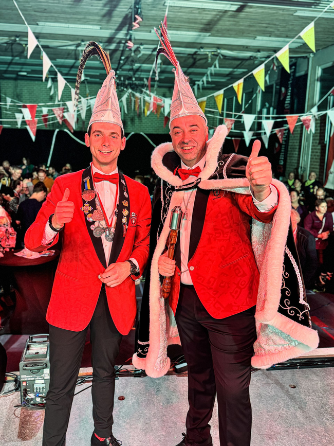 Twee mannen in kostuums poseren voor een foto en een van hen steekt zijn duim omhoog.