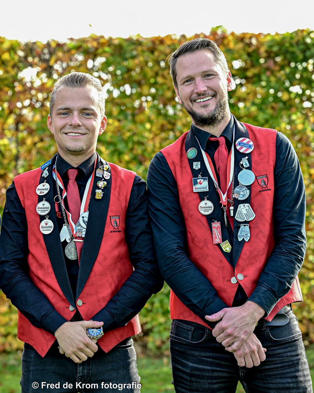 Drie mannen met medailles staan naast elkaar voor een heg.