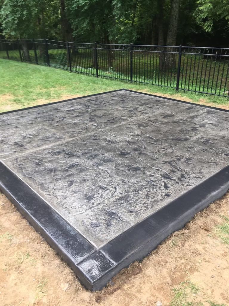 A square concrete slab with a black border is sitting in the middle of a yard next to a fence.