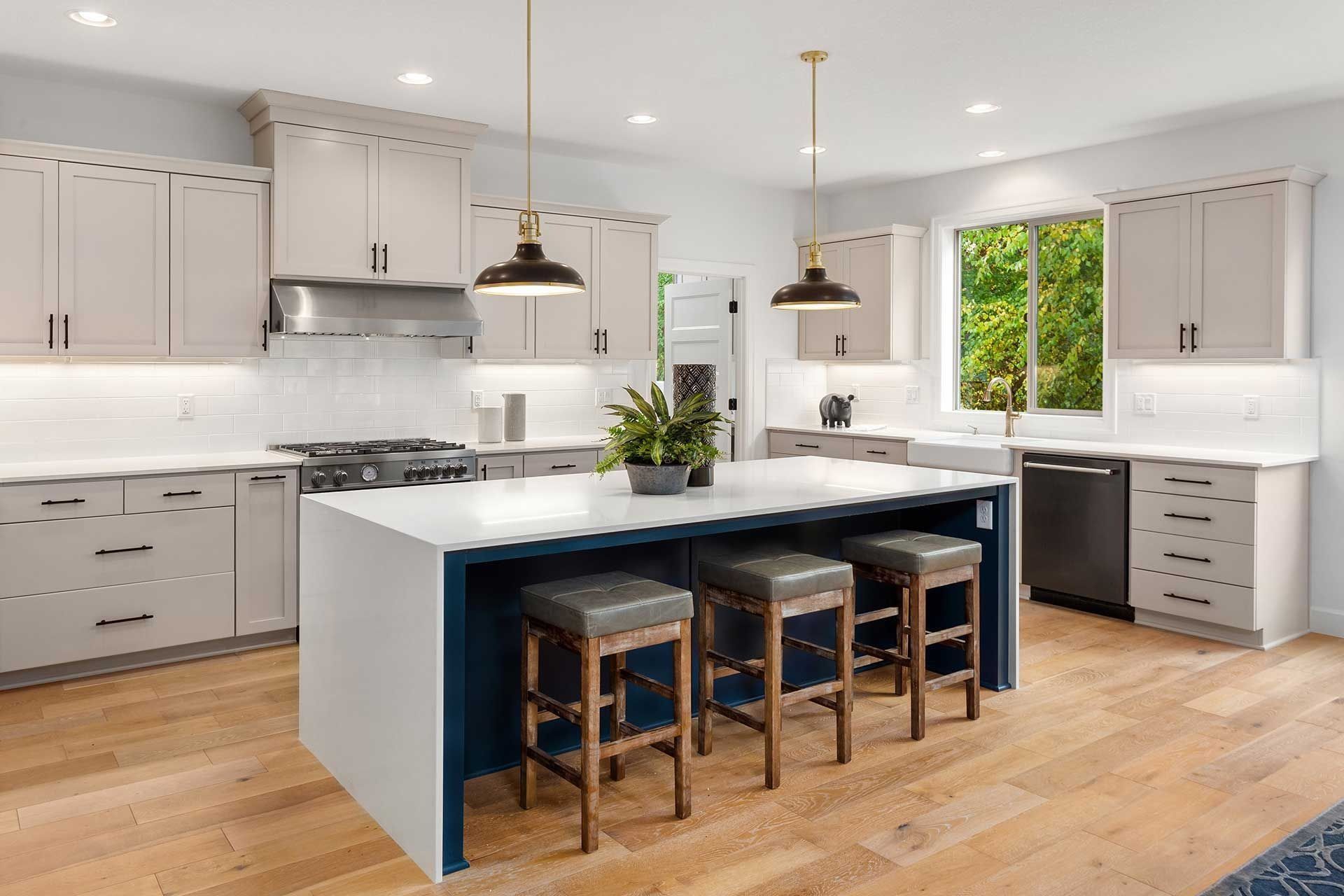 Modern kitchen with a white island, blue base, and three stools. Wooden floors and light beige cabinets.
