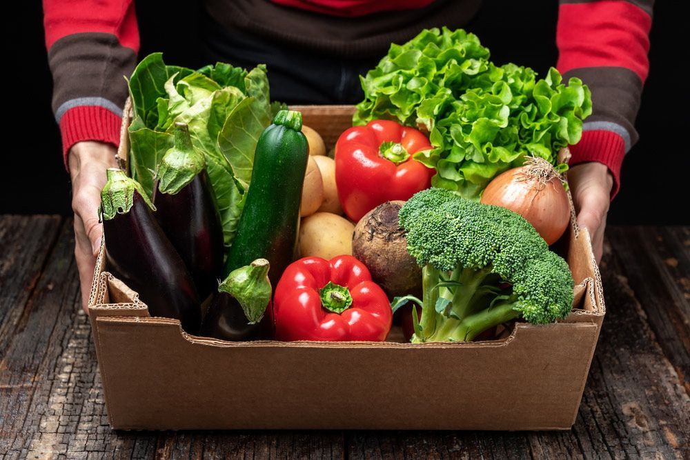 A Person Is Holding A Cardboard Box Filled With Vegetables — Remote Shopping Services In Edge Hill, QLD
