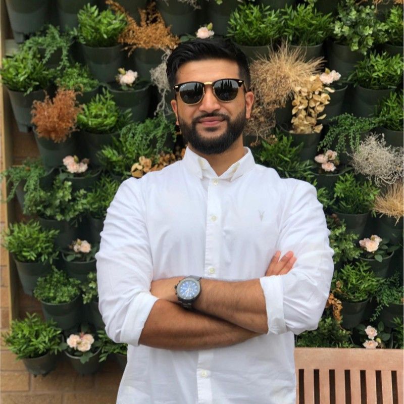 A man wearing sunglasses stands in front of a wall of potted plants