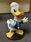 Een Donald Duck-figuurtje met open snavel, gekleed in een blauw matrozenpak, staand op een zwarte sokkel.