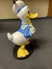 Een Donald Duck-figuurtje met open snavel, gekleed in een blauw matrozenpak, staand op een zwarte sokkel.