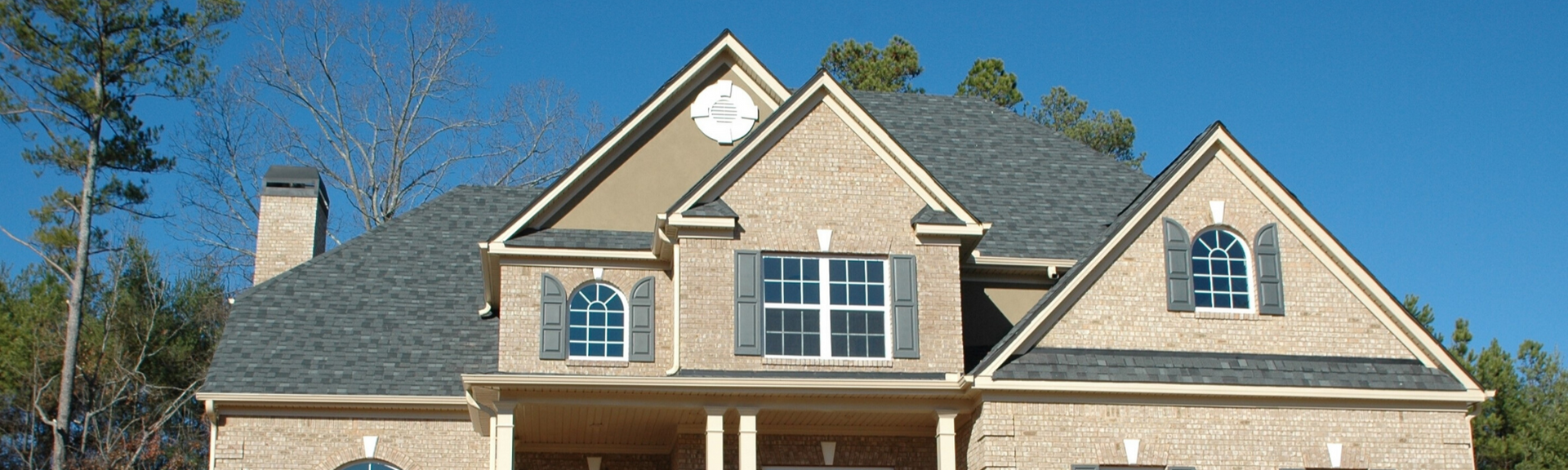 A brick two-story suburban house with a gray shingled roof, multiple gables, and dark shutters under a clear blue sky.