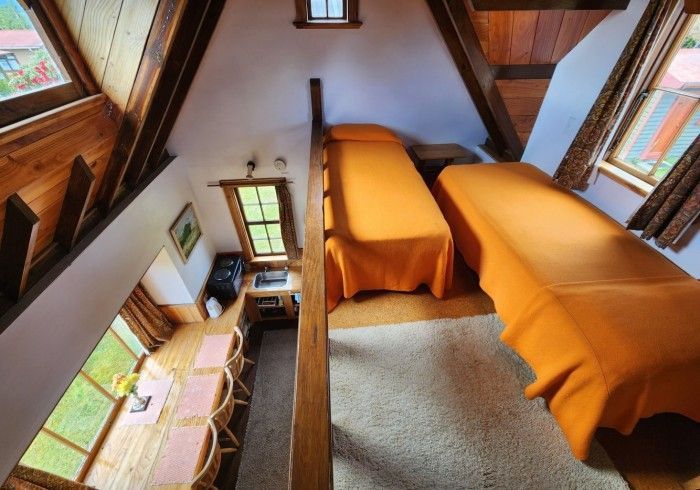 Loft bedroom with two beds, orange covers, exposed wooden beams, and a dining table below.