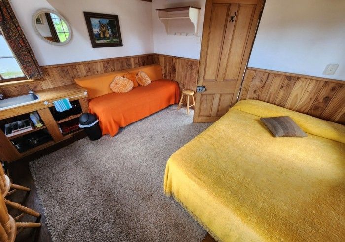 Cozy cabin interior with bed, sofa, kitchenette, wood paneling, and a small round mirror.