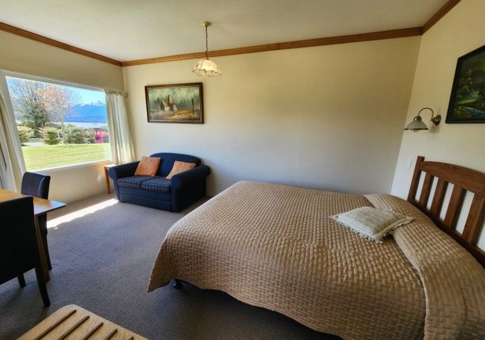 Bedroom with bed, small sofa, window overlooking a lawn and lake. Beige walls, brown trim.