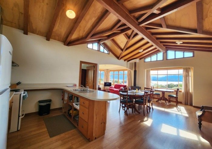 Open-plan living space with kitchen, dining area, and living room featuring wooden beams, windows, and water views.