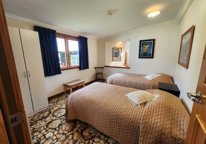 Two beds in a room, brown comforters, a window, blue curtains, a wardrobe, and art on the wall.