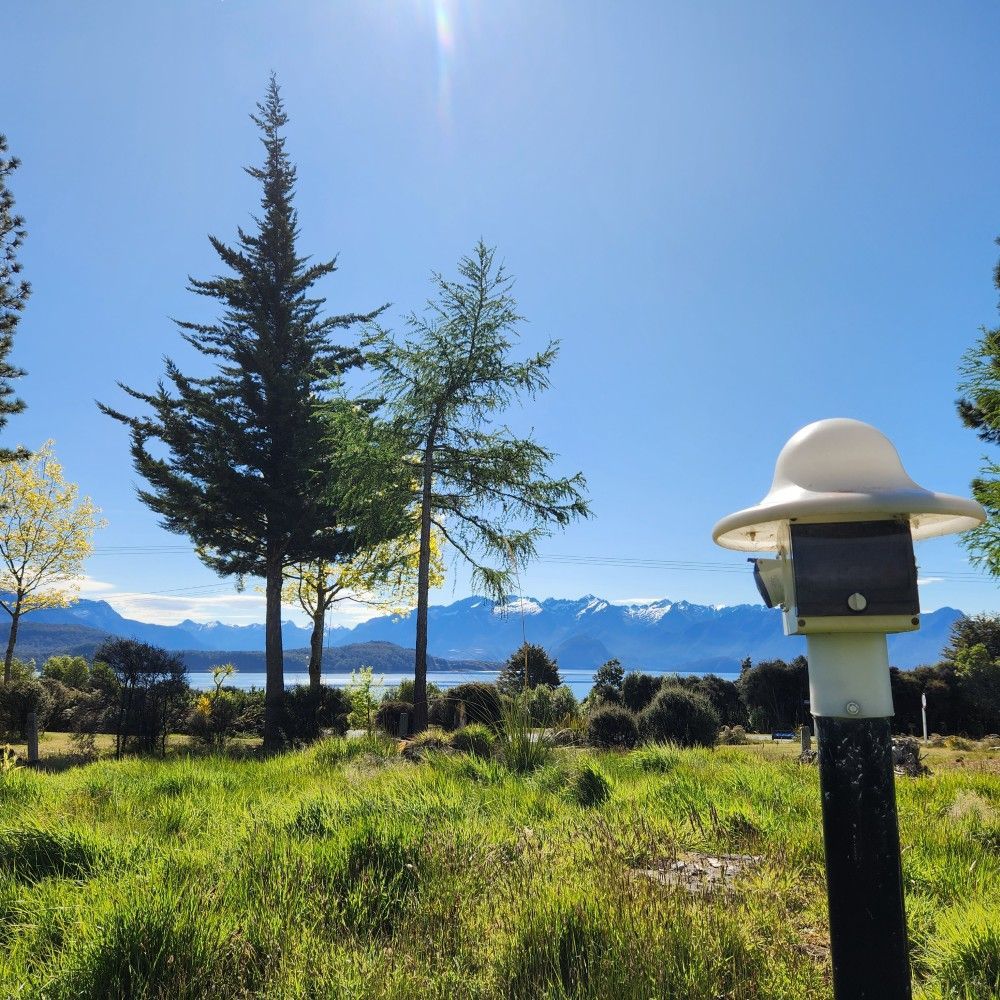 Sunny landscape with green grass, trees, lake, and snow-capped mountains under a blue sky. A white lamp post is in the foreground.