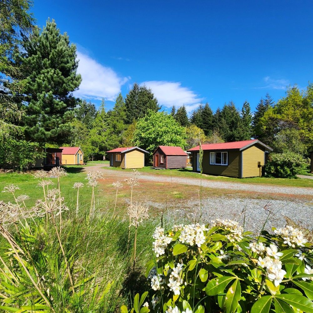 Small cabins with red roofs on a green lawn, under a blue sky, surrounded by trees.