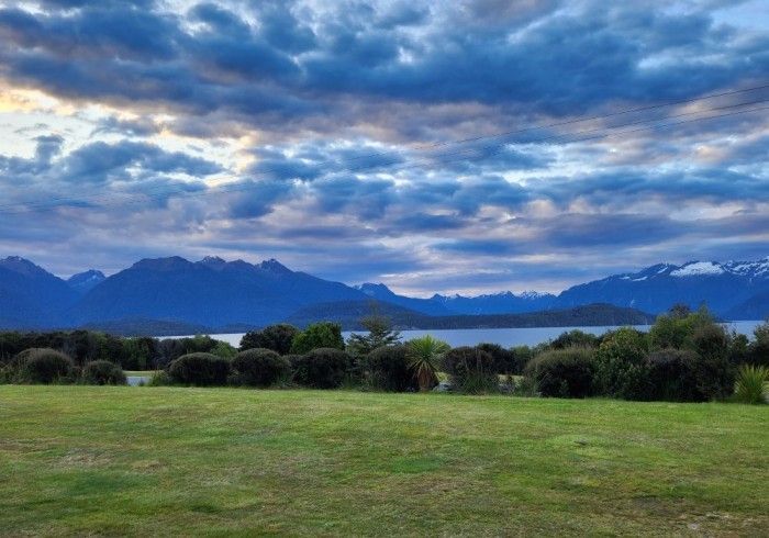 Green lawn with bushes, calm lake, and mountains under a cloudy, blue and orange sky.