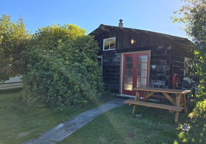 Weathered dark wooden cabin with red doors and a picnic table. Lush green bushes and grass surround the building.