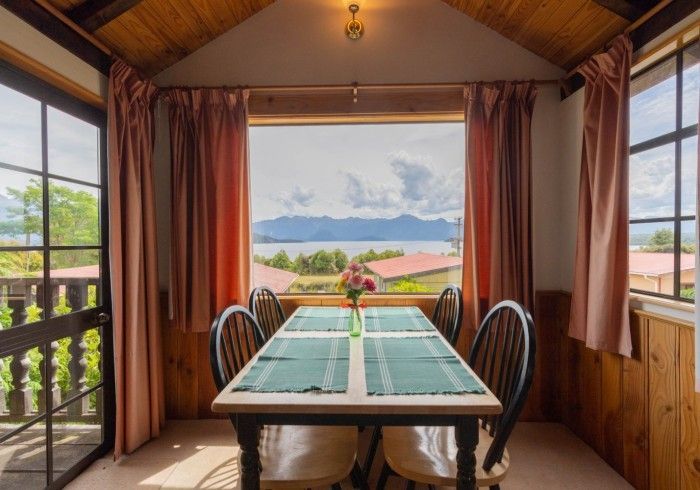 Dining room with table set for six, large window overlooking water and homes. Pink curtains, wooden paneling.
