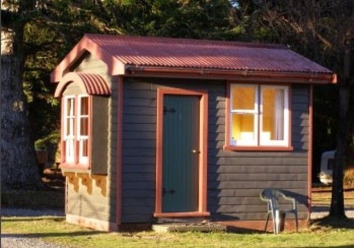 Small, gray wooden shed with a red corrugated metal roof, door, and two windows in a grassy setting.