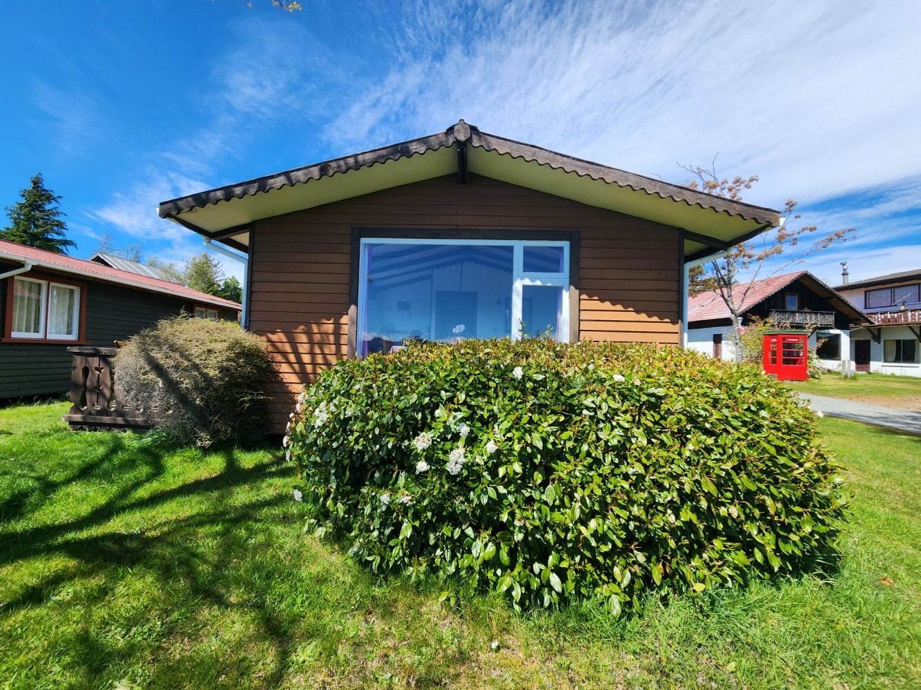Brown cabin with large window and trimmed green bush in front, blue sky overhead.