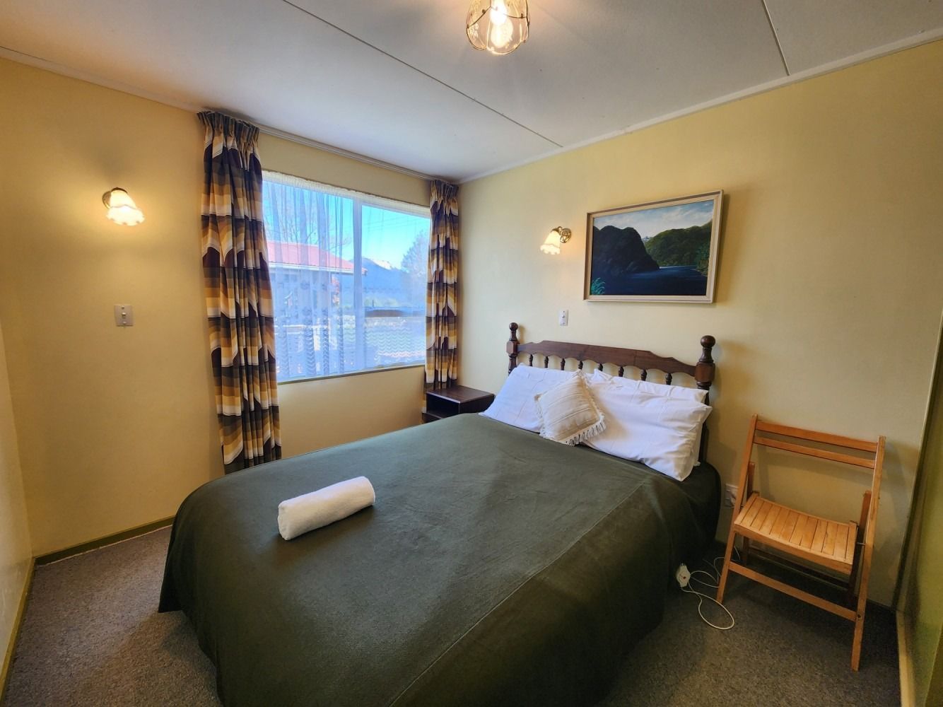 Bedroom with a bed covered in green, a window, artwork, and a wooden chair.