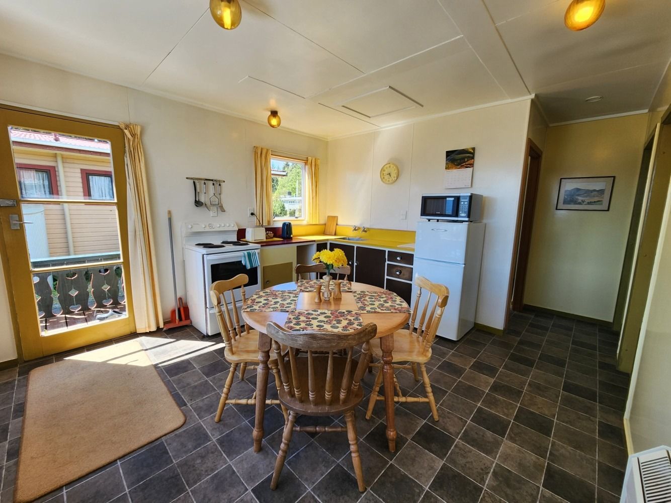 Kitchen with a round wooden table and chairs, sunlight streaming in, yellow and dark-tiled floor.