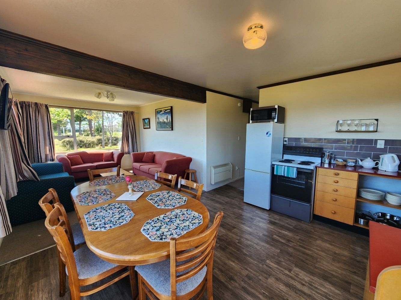 Open-plan living space with dining table, sofas, and kitchen appliances. Wooden floors, tan walls, and natural light.